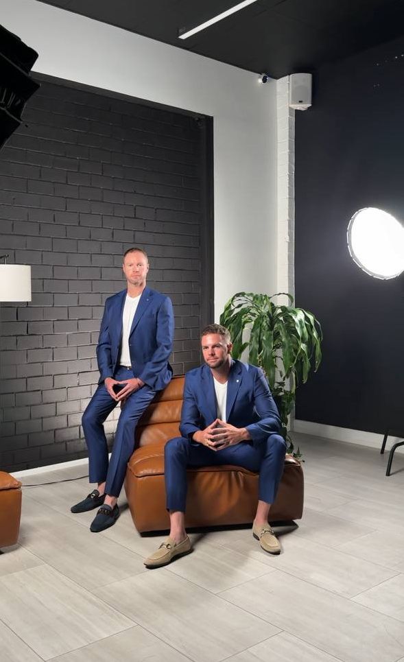 Two people in matching blue suits posing on brown leather furniture in modern studio setting.