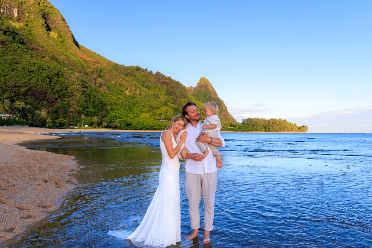 Two people in light colored attire embrace on a scenic beach with mountains and blue ocean water in Hawaii at sunset.