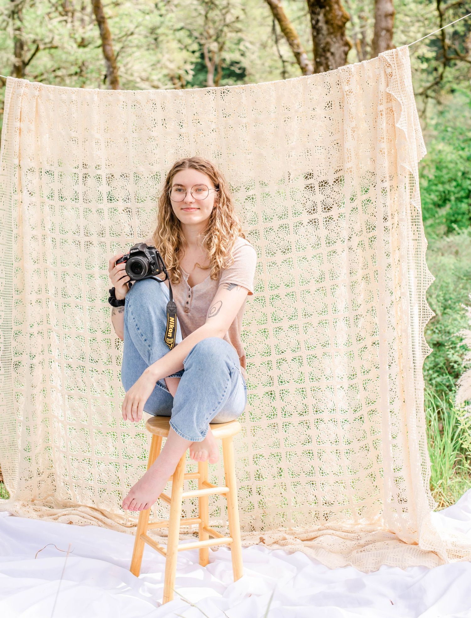 Person in beige top and jeans poses with camera against decorative textile backdrop in garden setting.