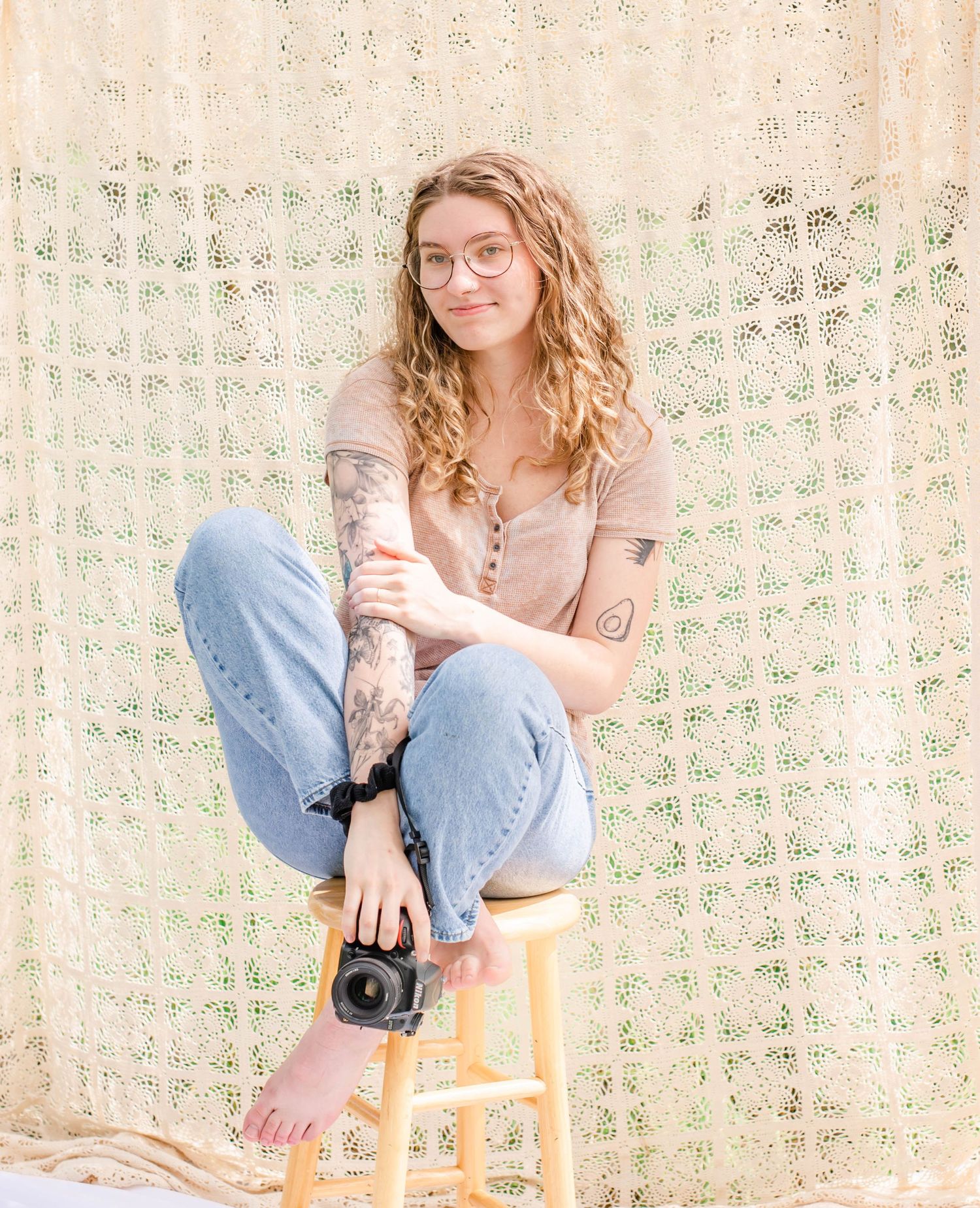 Photographer sits casually on wooden stool against macrame backdrop holding camera in natural outdoor setting.