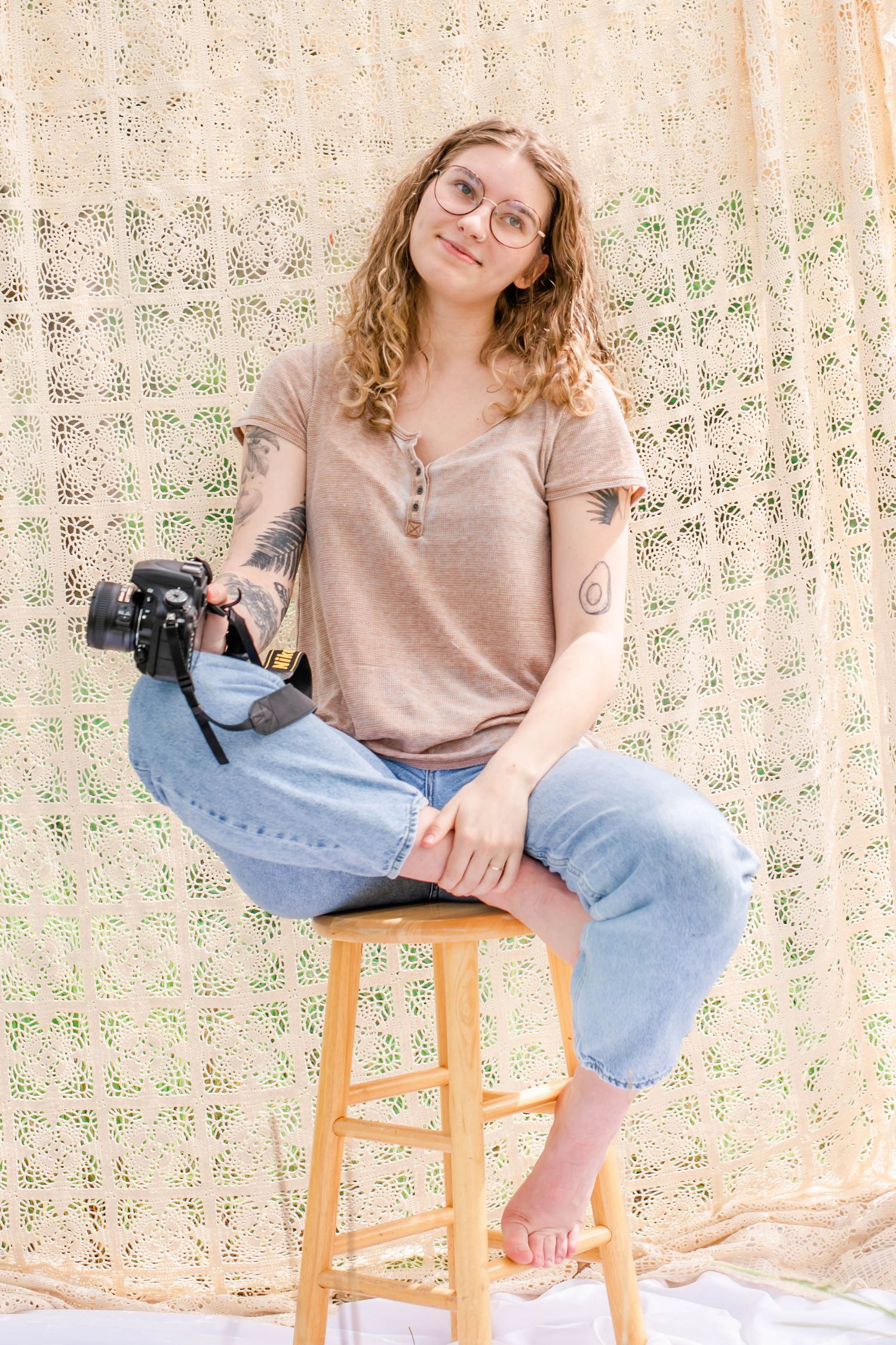 Woman sits casually on wooden stool against vintage lace curtain in natural lighting.
