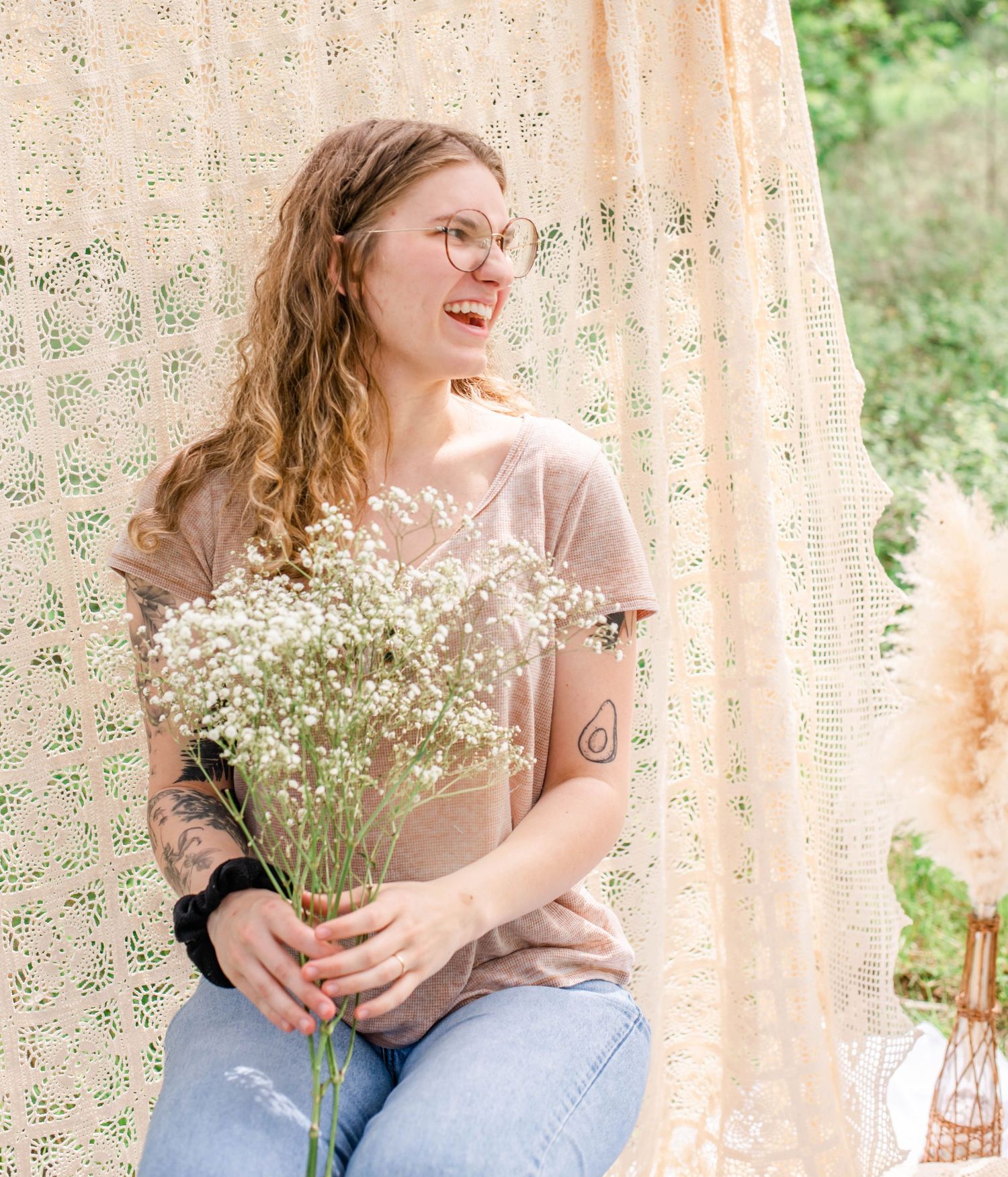 Woman holding baby's breath flowers poses against cream lace curtain in outdoor setting.