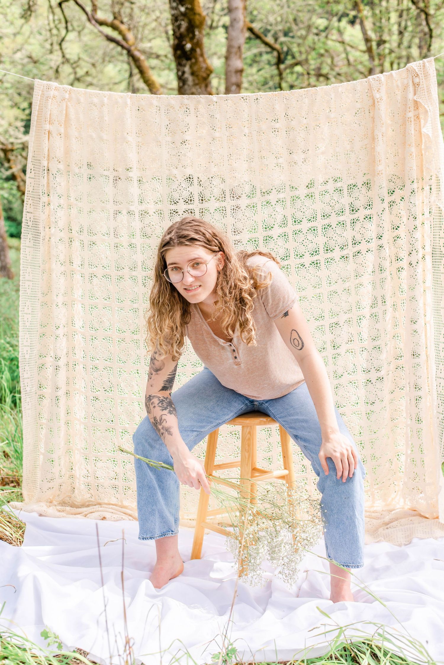 Woman in casual wear poses on wooden stool with white fabric and lace curtain backdrop.