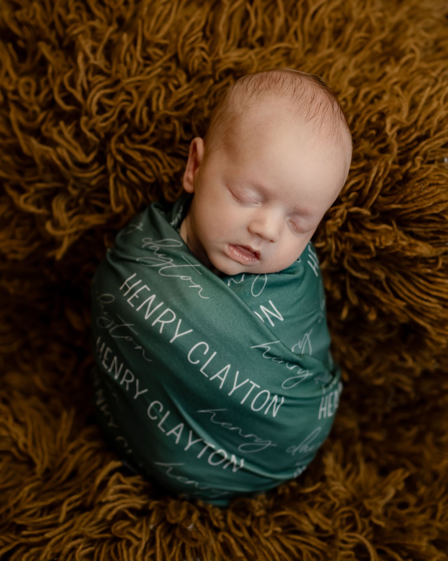 Sleeping newborn swaddled in a green personalized blanket on textured brown fabric.