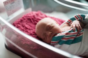 Newborn baby resting in hospital bassinet with pink and teal blankets.