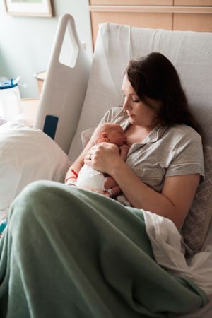 Mother rests with newborn baby under green blanket in hospital bed.