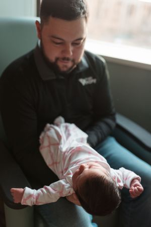 A tender moment between parent and newborn baby in a black shirt by a window.