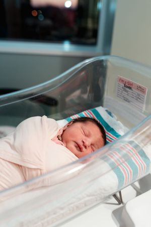 Sleeping newborn wrapped in white blanket resting in clear hospital bassinet with striped bedding.