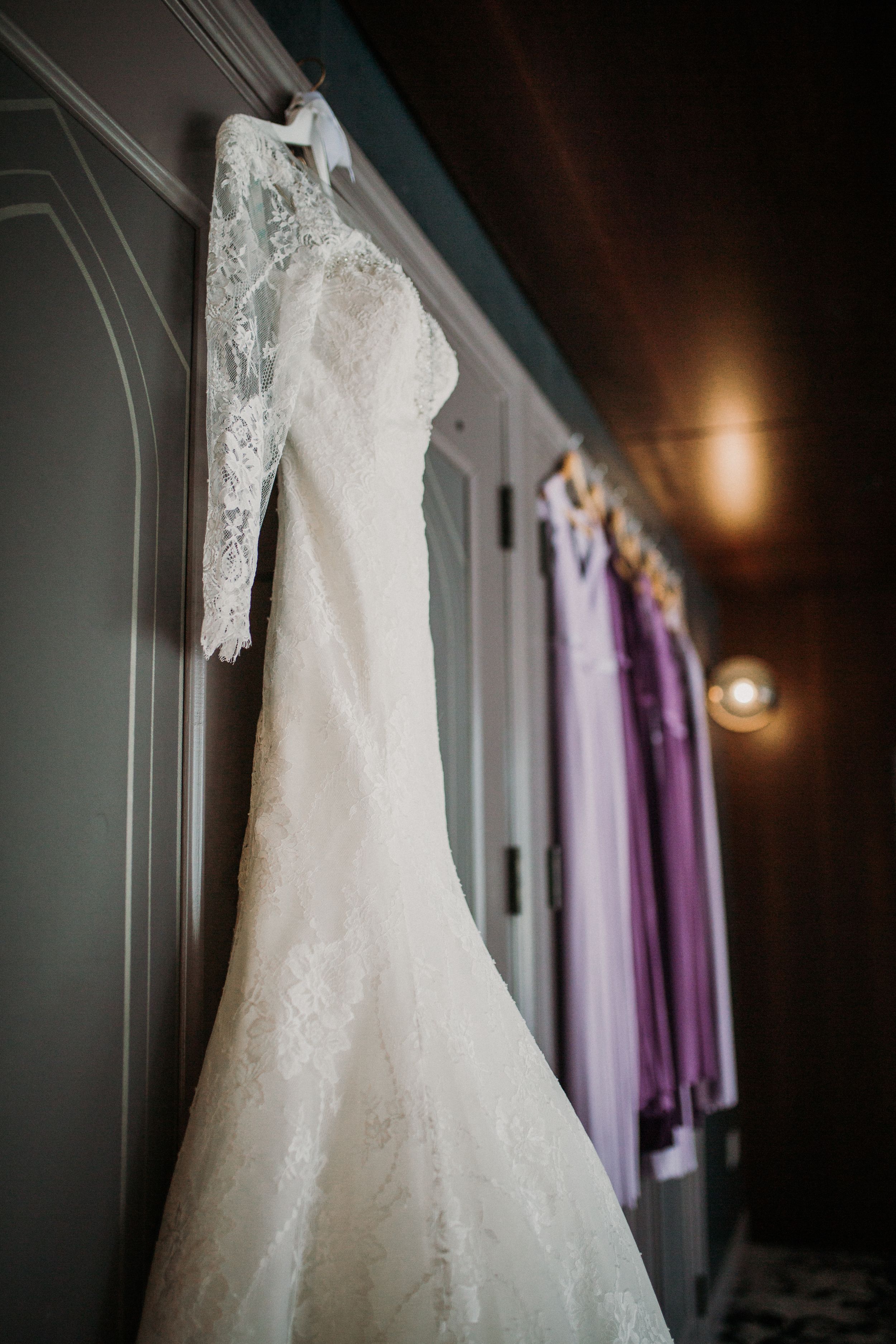 A delicate lace wedding dress hangs beside purple bridesmaid dresses in dim lighting.