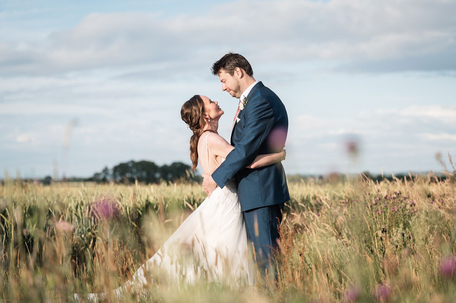 Romantic embrace between newlyweds in field of tall grass under cloudy sky.