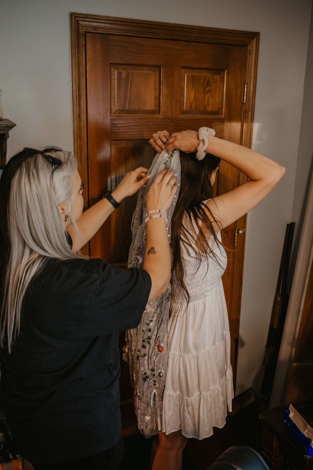 Bridesmaid helps bride-to-be put on embroidered shawl in front of wooden door.