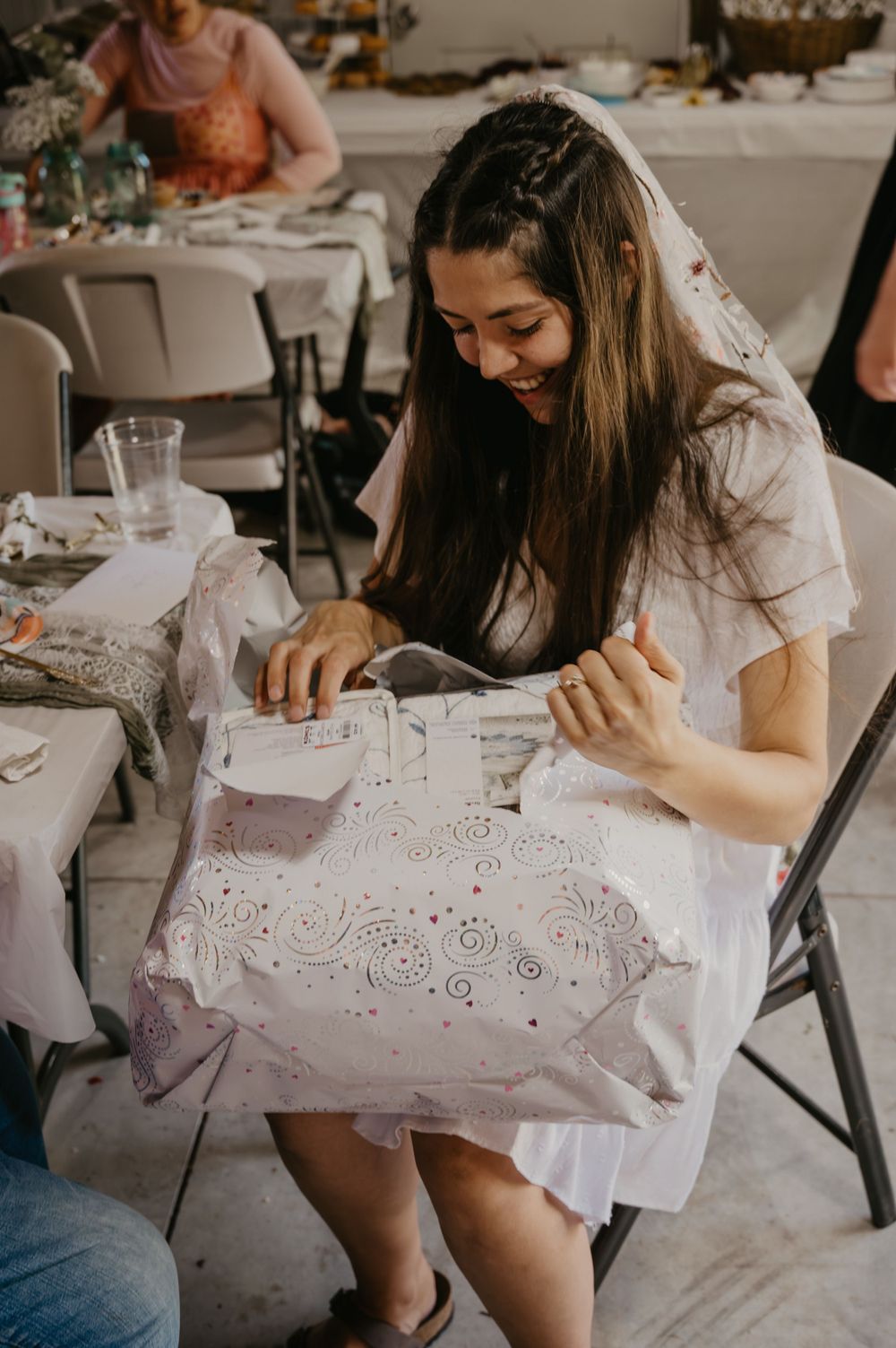 Smiling bride-to-be opening a gift at her bridal shower seated at a decorated table.