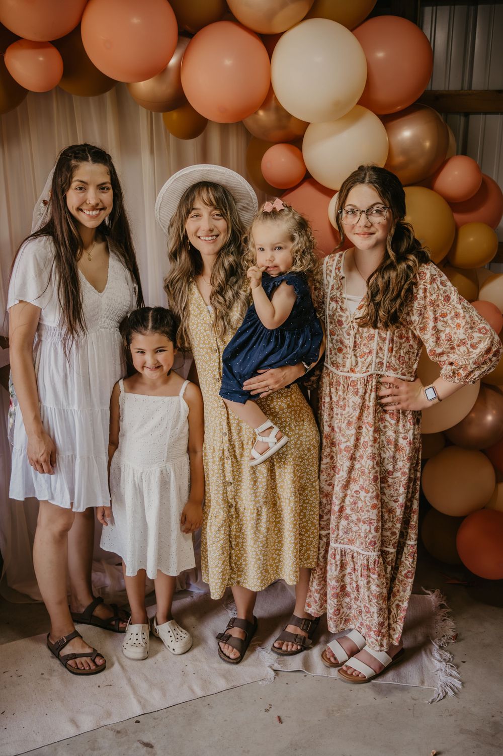 Group of four females posing in front of a boho peach and gold balloon arch at a bridal shower.