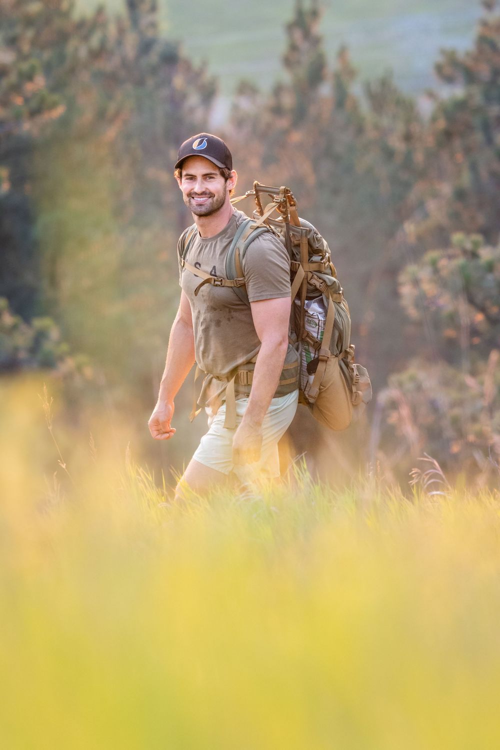 A hiker with a backpack smiles while walking through a sunlit meadow during golden hour.