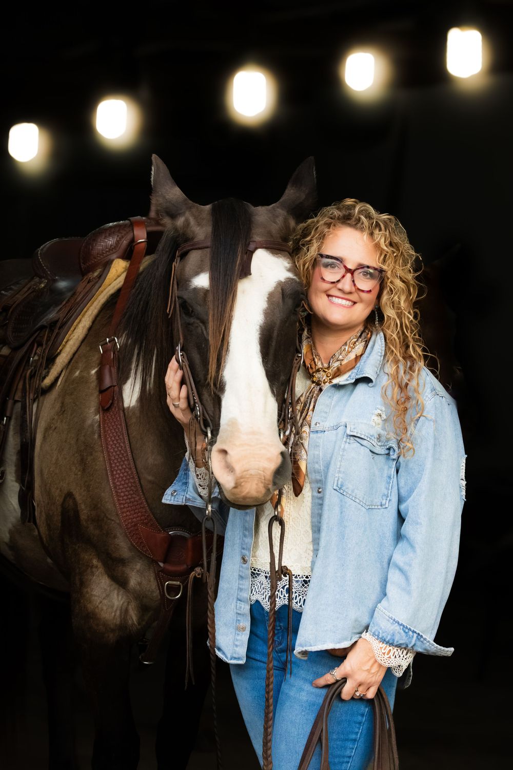 A person in a denim jacket poses with a brown horse at night with string lights in the background.