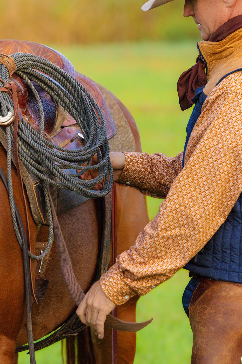 Close-up of someone wearing a yellow polka dot shirt and navy vest adjusting ropes on a brown leather western saddle.