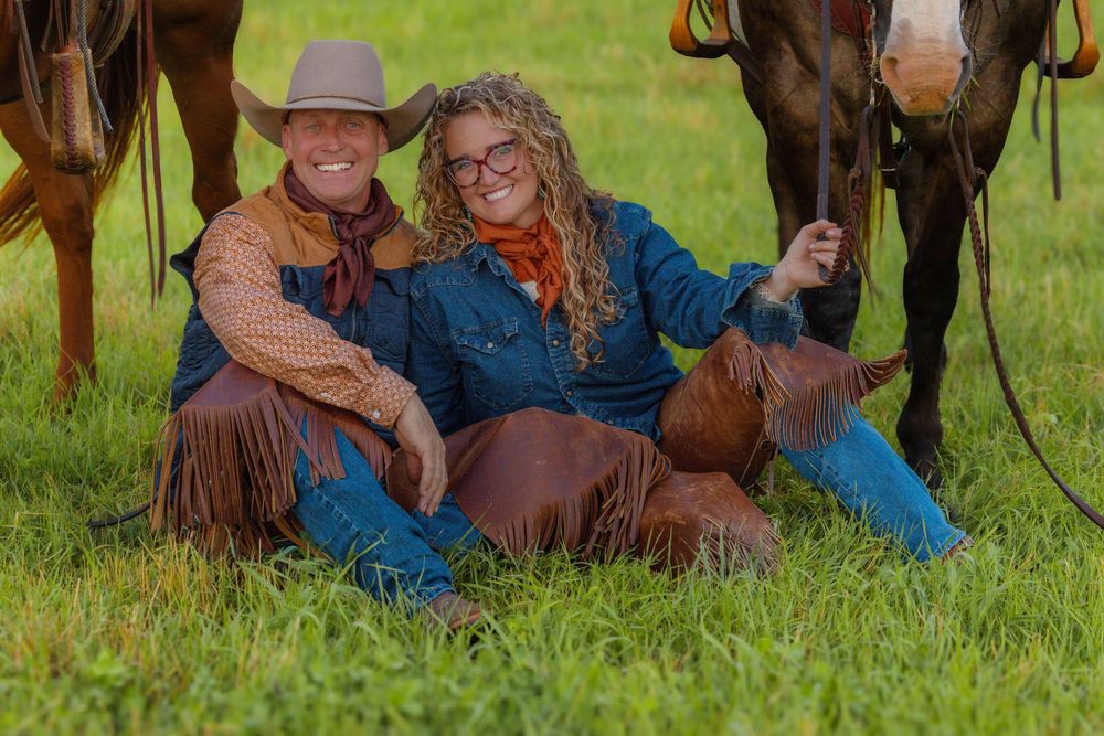 Cowboy and companion resting in green grass field next to horses during western photoshoot.