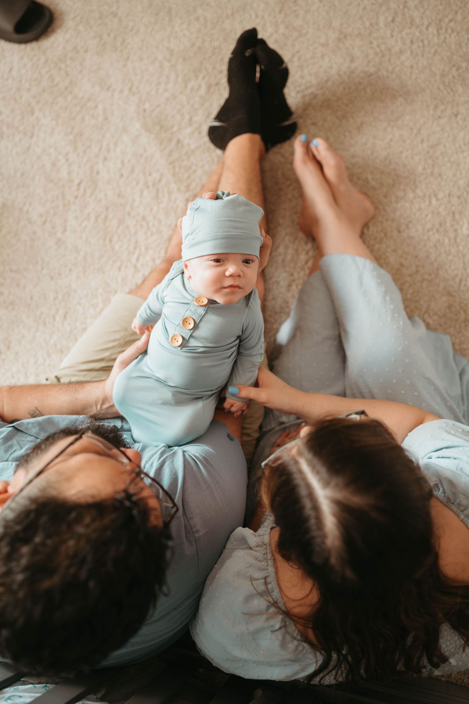New parents lying on floor bonding with their newborn baby dressed in sage green outfit and matching hat.