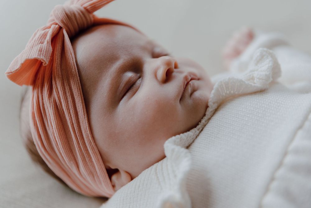 A sleeping baby wearing a pink headband lies peacefully on white bedding.
