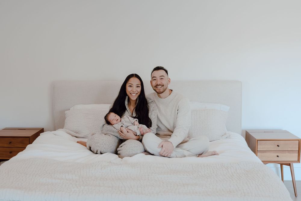 A family cuddles together under white bedding in a minimalist bedroom setting.