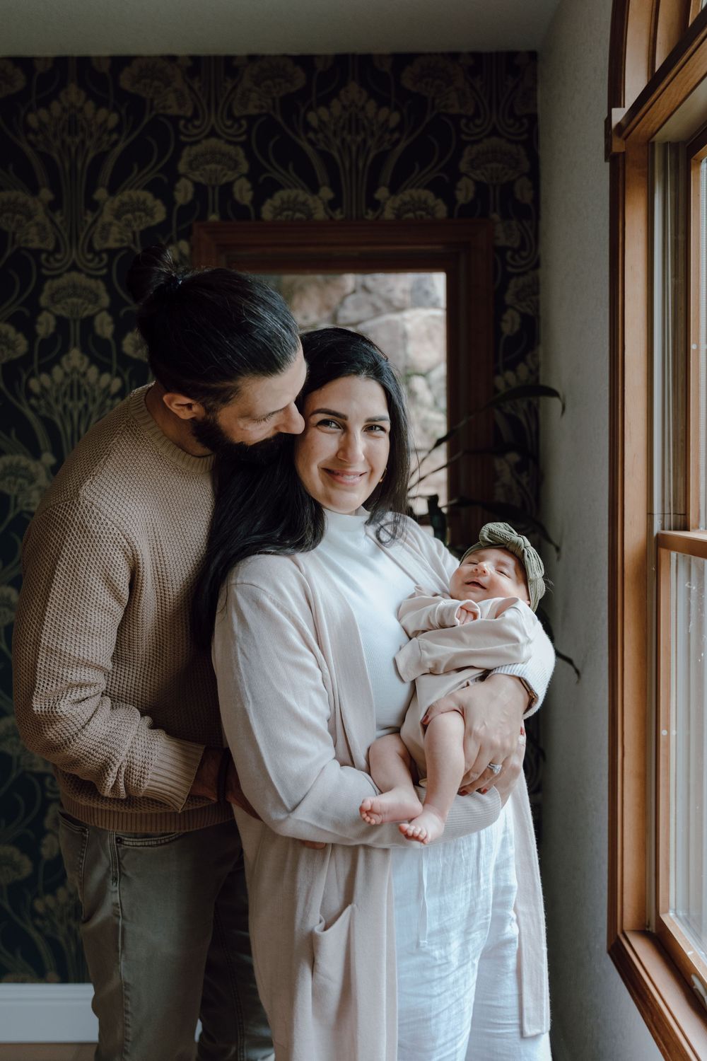 A tender moment captured by a window with soft natural light and elegant damask wallpaper.