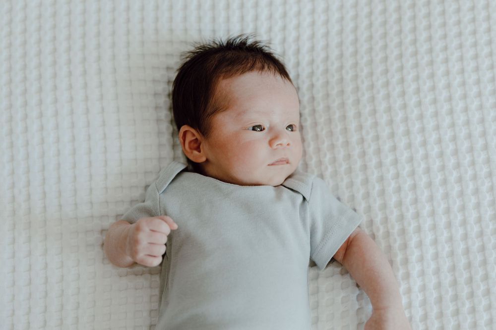 Baby wearing light gray onesie lies on textured white blanket looking to the side.