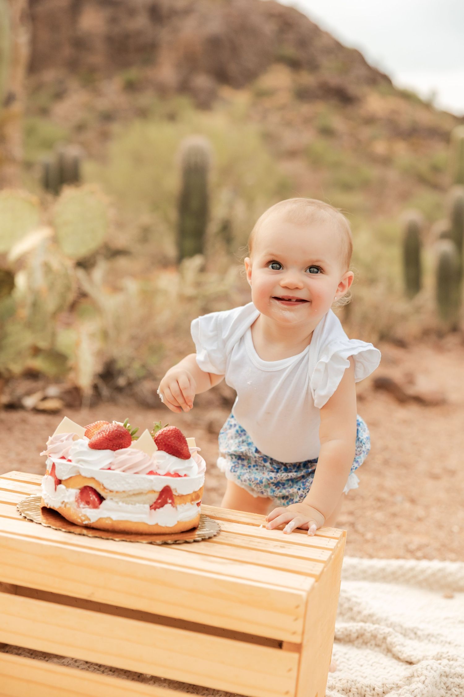 A baby in white clothing smiles brightly while reaching for a strawberry dessert on a wooden table in a desert setting.
