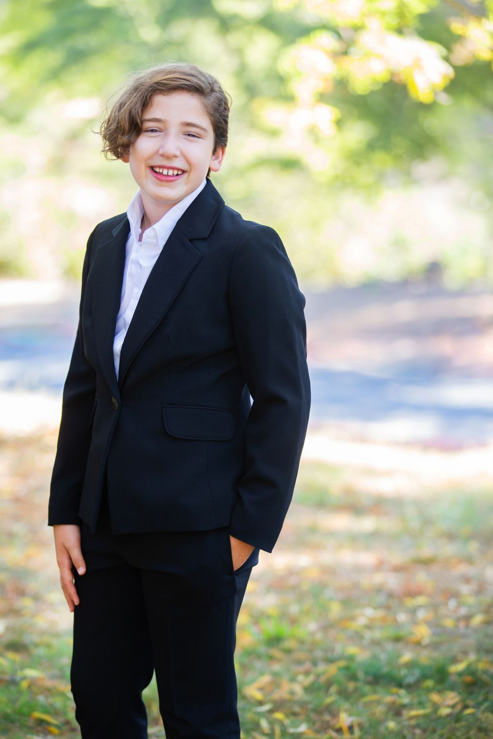 Young person in a dark suit and white shirt smiling outdoors on an autumn day with blurred natural background.