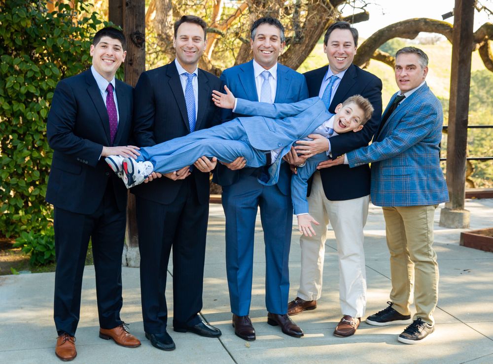 Group in blue suits and formal wear playfully lift and support one member during an outdoor photo session.