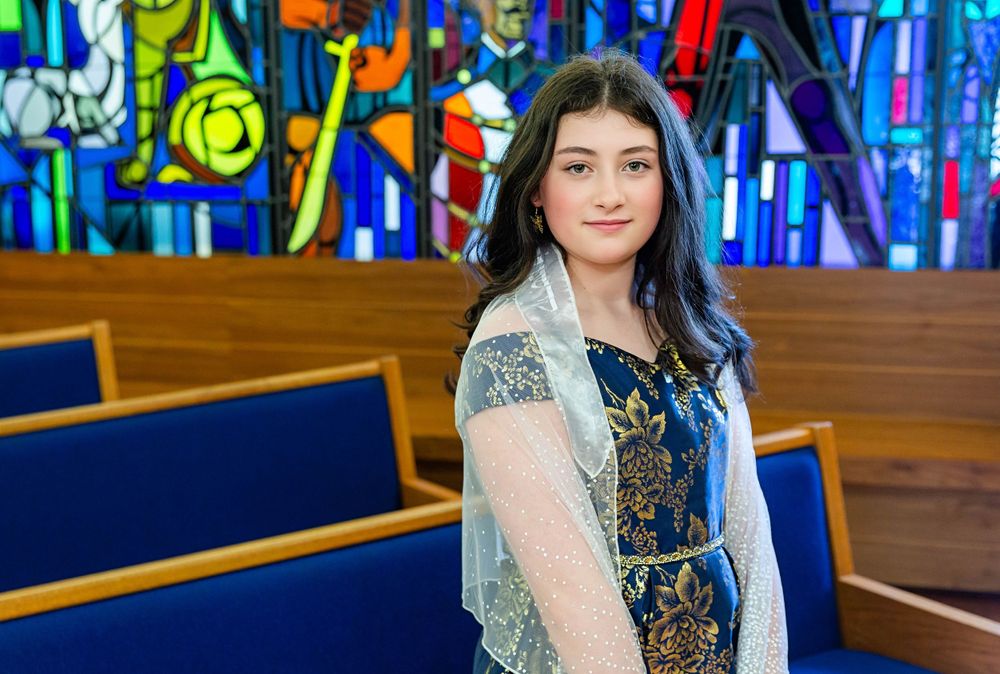 Colorful stained glass church window provides vibrant backdrop for person in elegant dress with lace overlay.