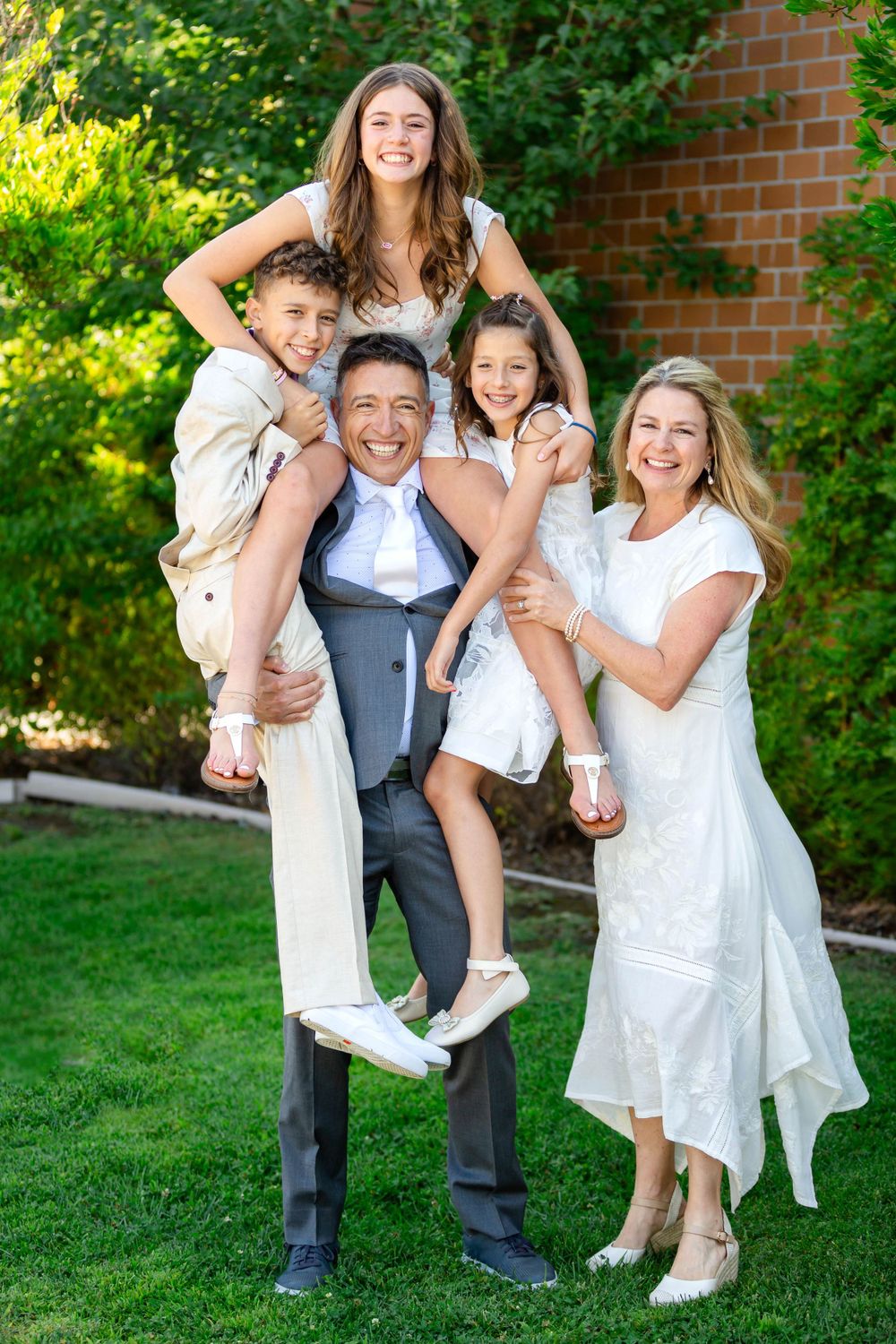 A joyful family stands together outdoors in front of a brick wall and greenery on a sunny day.