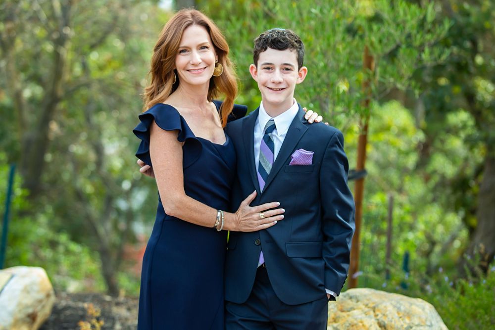 A person in a navy dress poses with a young person wearing a navy suit and striped tie in an outdoor garden setting.