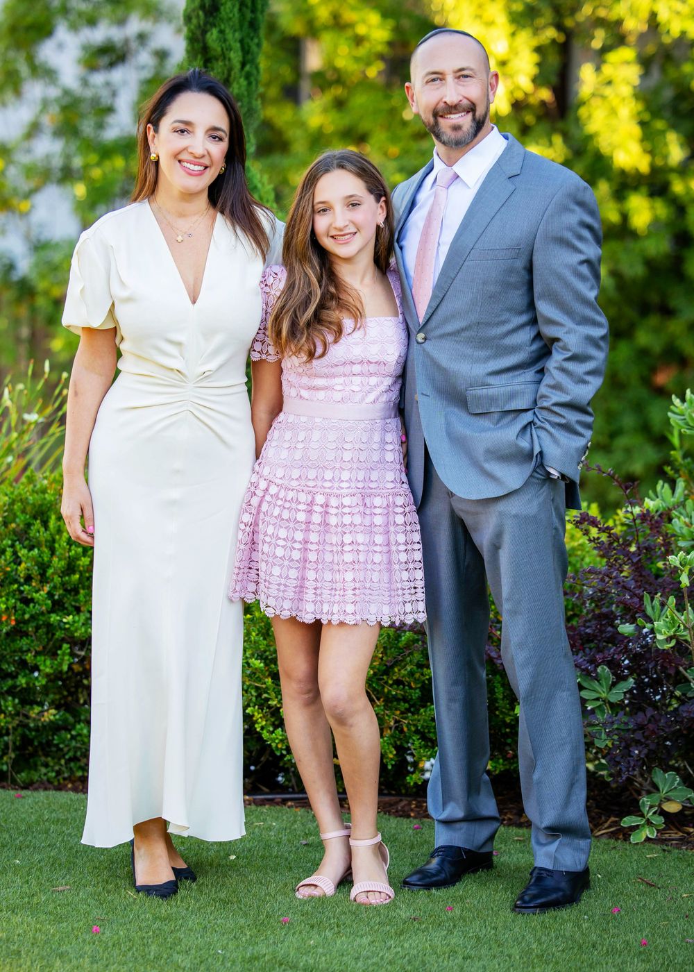 A family of three poses together outdoors wearing formal attire including a white dress, pink dress and gray suit.