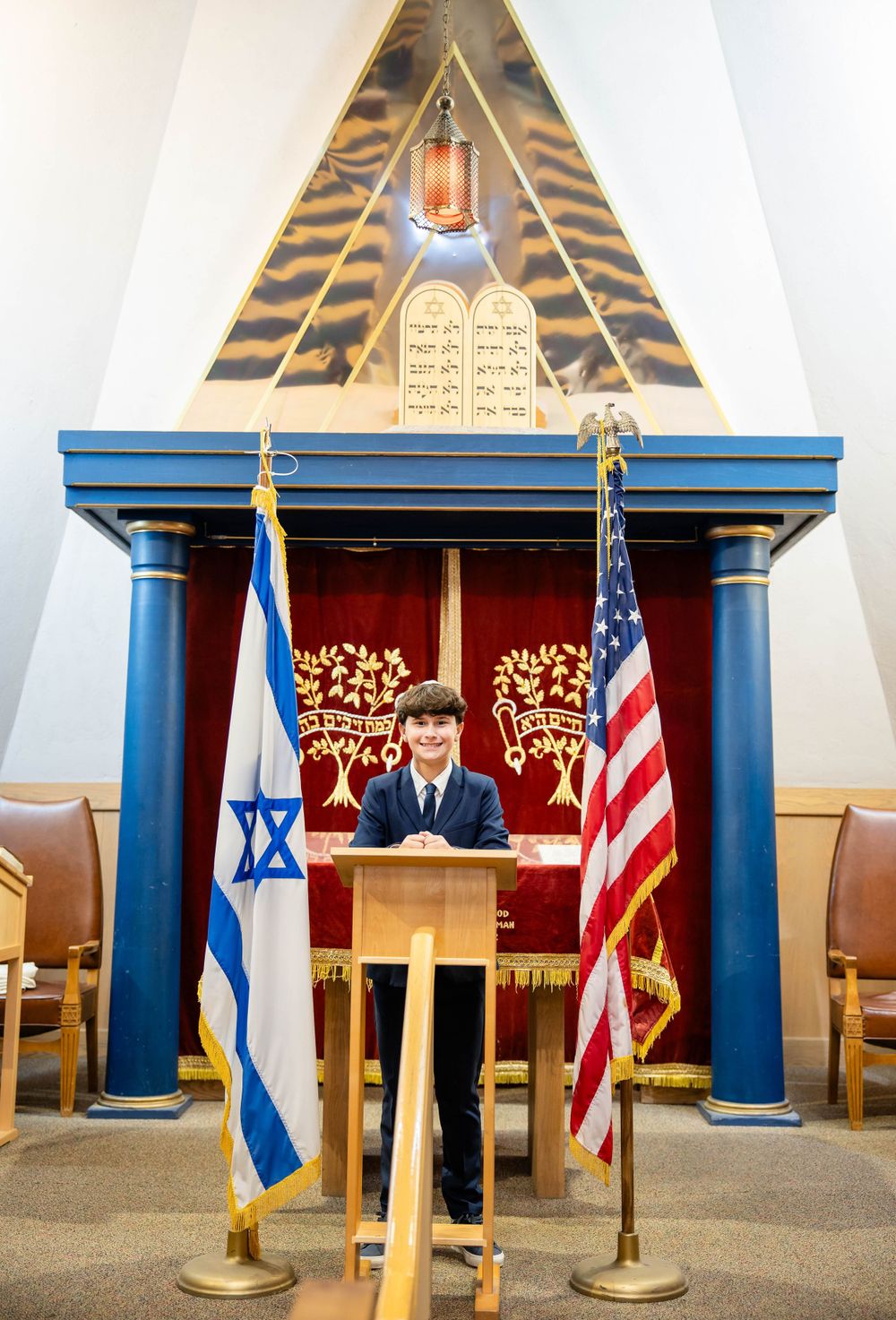 A synagogue podium features two flags - Israeli and American, with decorative pillars and Torah symbols in the background.