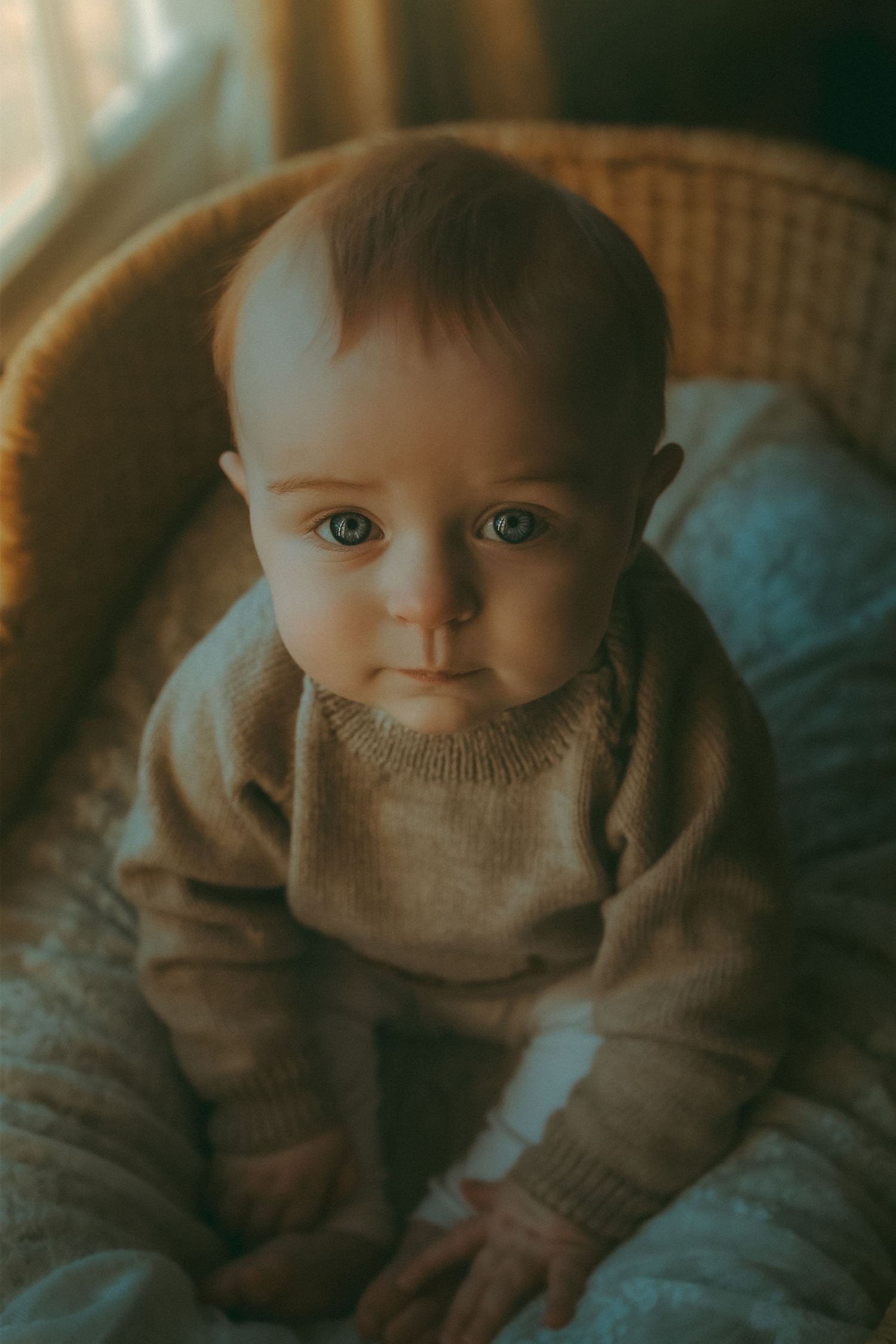 Bright-eyed baby sitting in wicker chair – cozy natural light baby photography