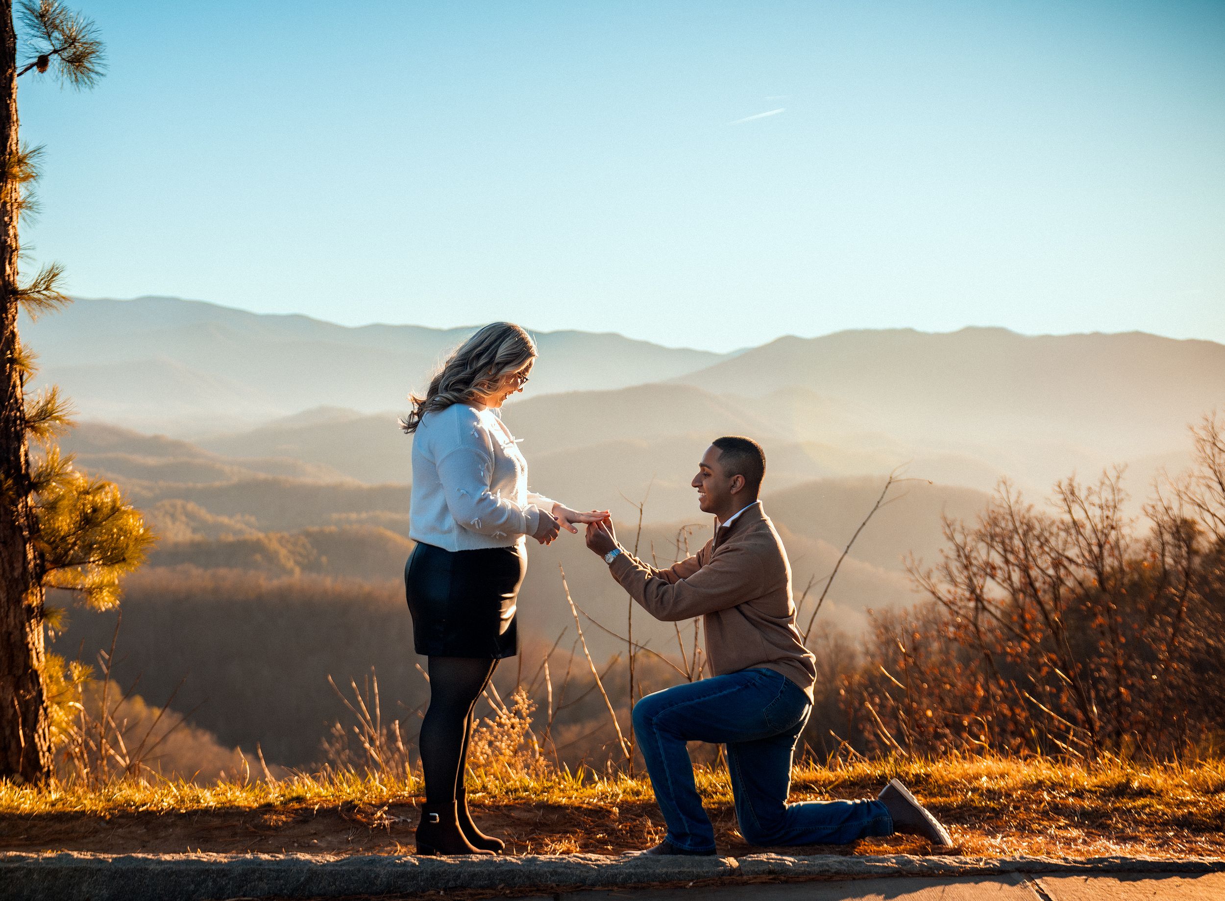 A couple shares a romantic proposal moment at sunset on a mountain overlook with golden sunlight and hazy peaks.