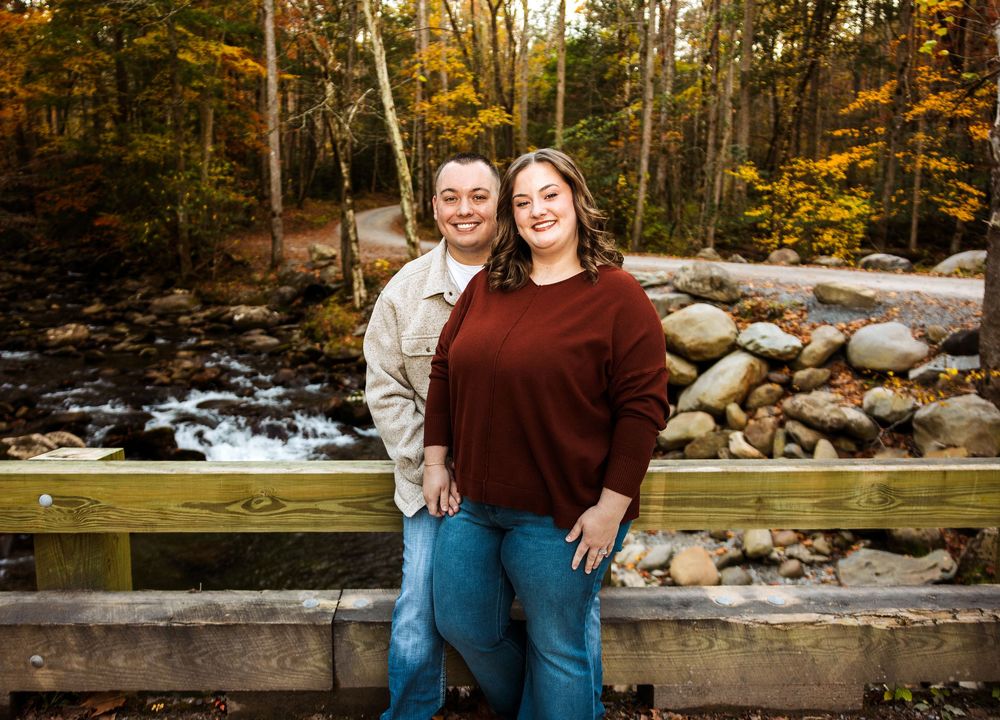 Two people stand on a wooden bridge over a rocky stream during autumn with colorful fall foliage in the background.