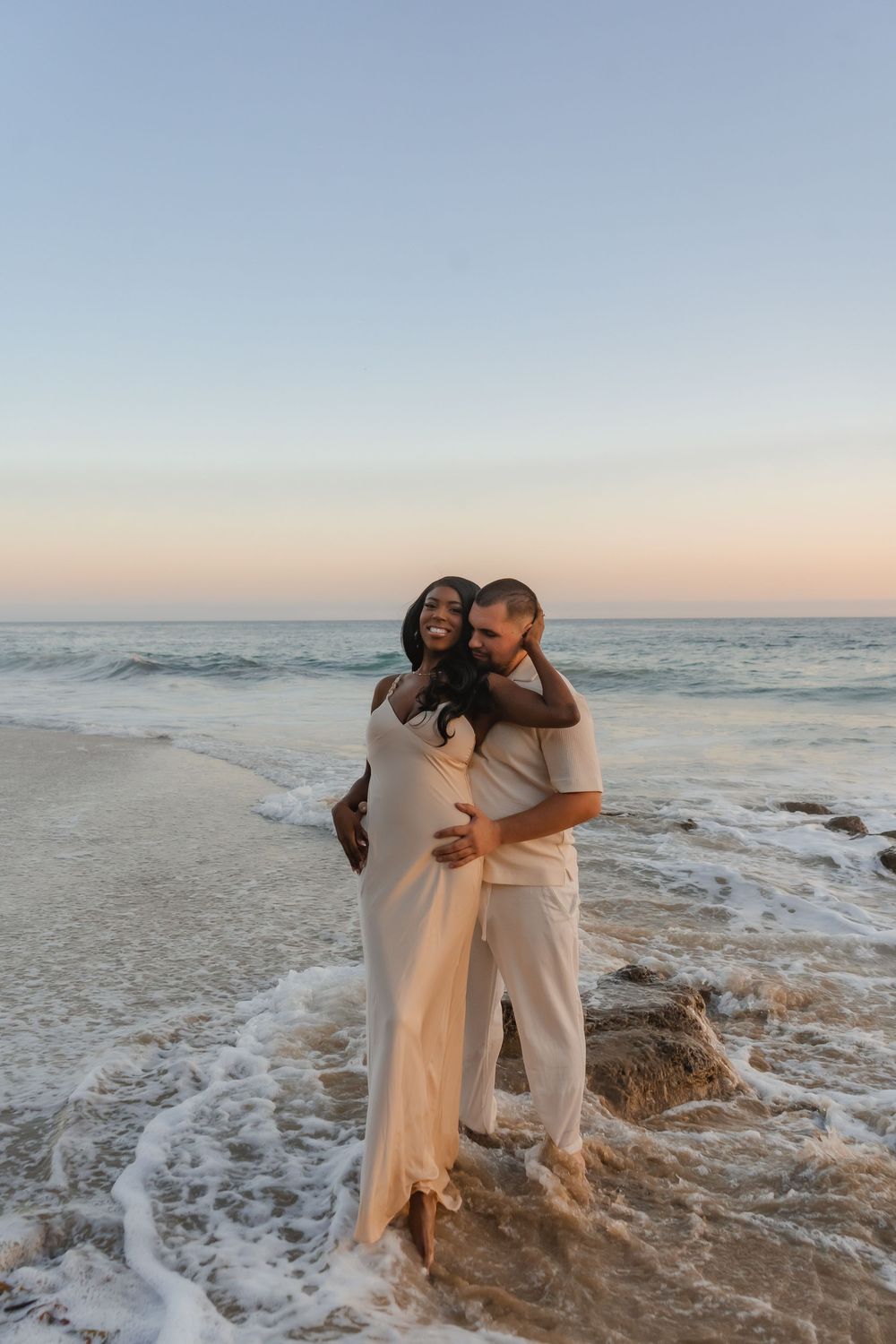 Two people embrace in flowing outfits at sunset on a beach as gentle waves roll in behind them creating a romantic moment.