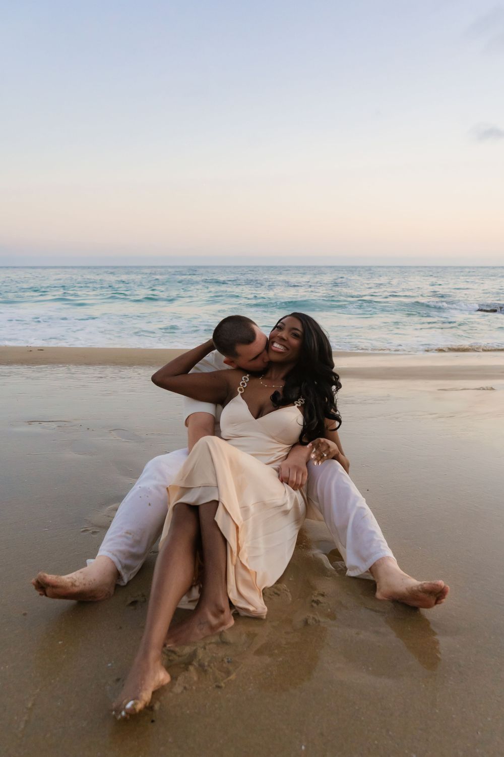 A romantic couple embraces on the beach at sunset while sitting in the sand with ocean waves in the background.