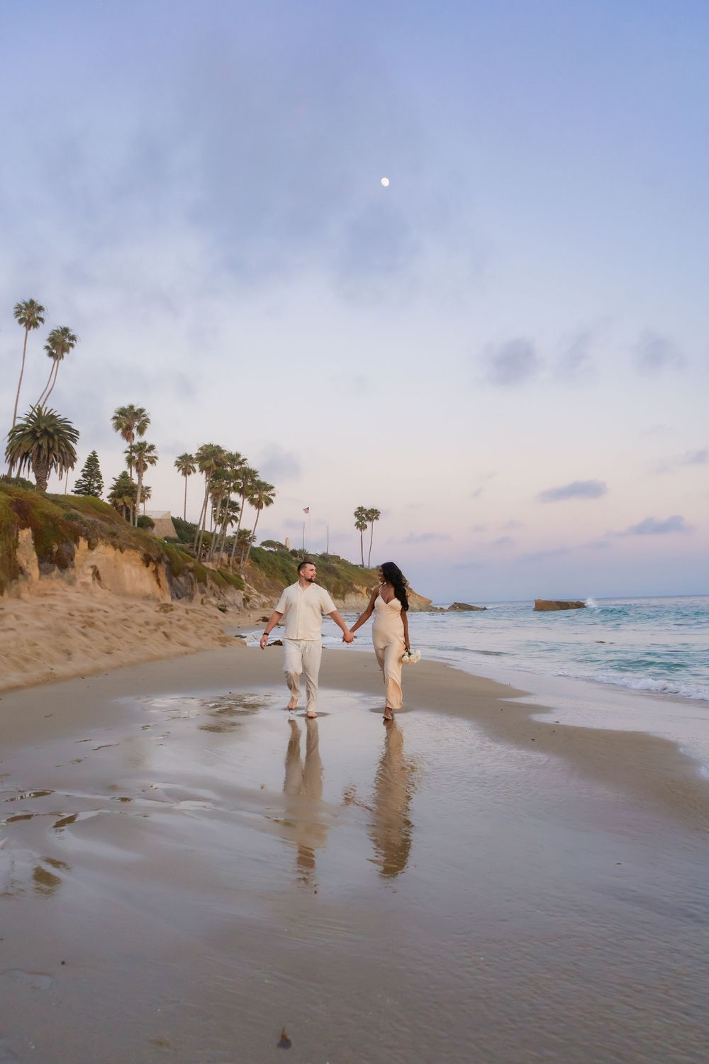 A couple walking along a misty beach shoreline at sunset with palm trees in the background creating a romantic mood.