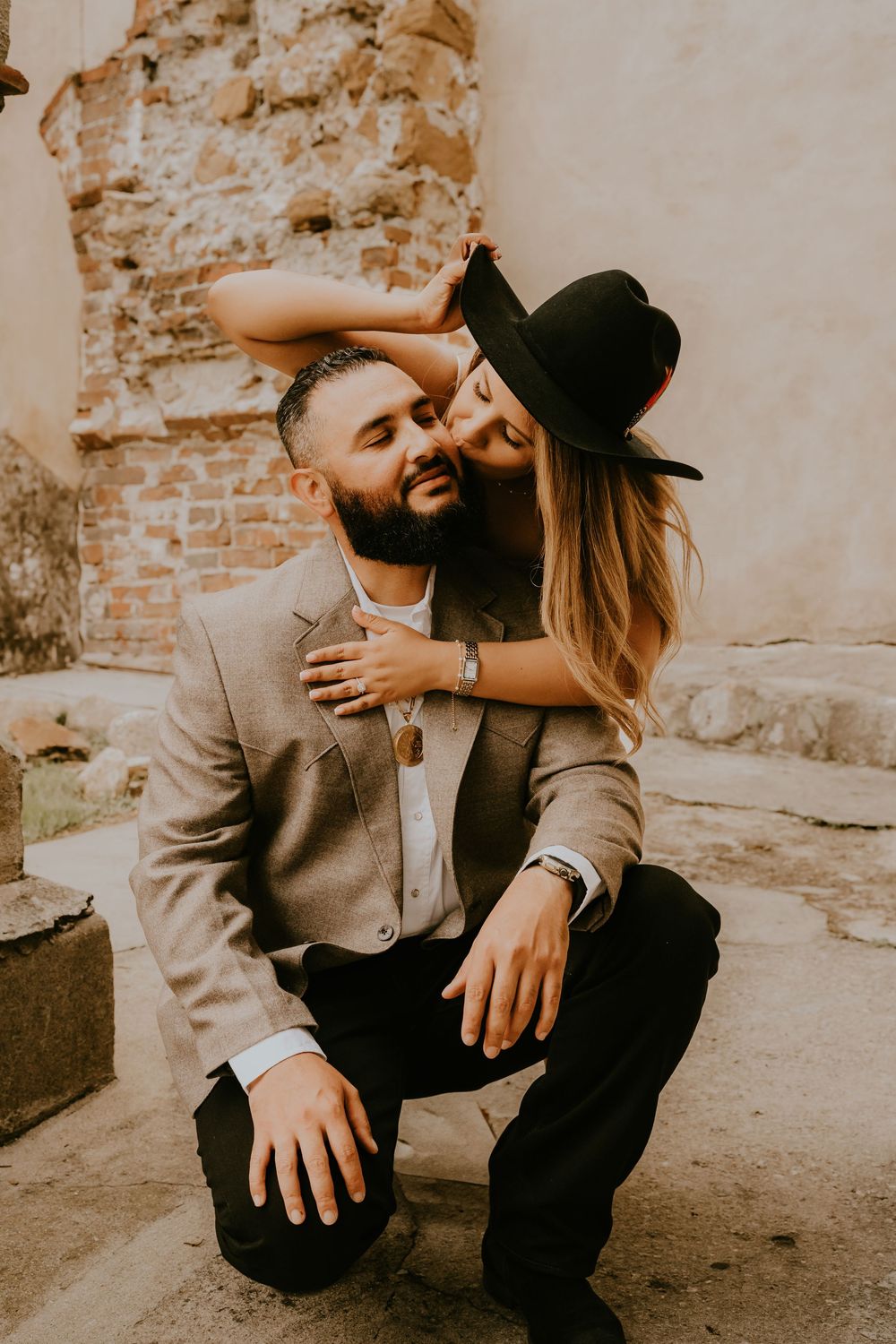 A romantic couple embraces in warm sepia tones against a weathered brick wall as she leans in wearing a black hat.