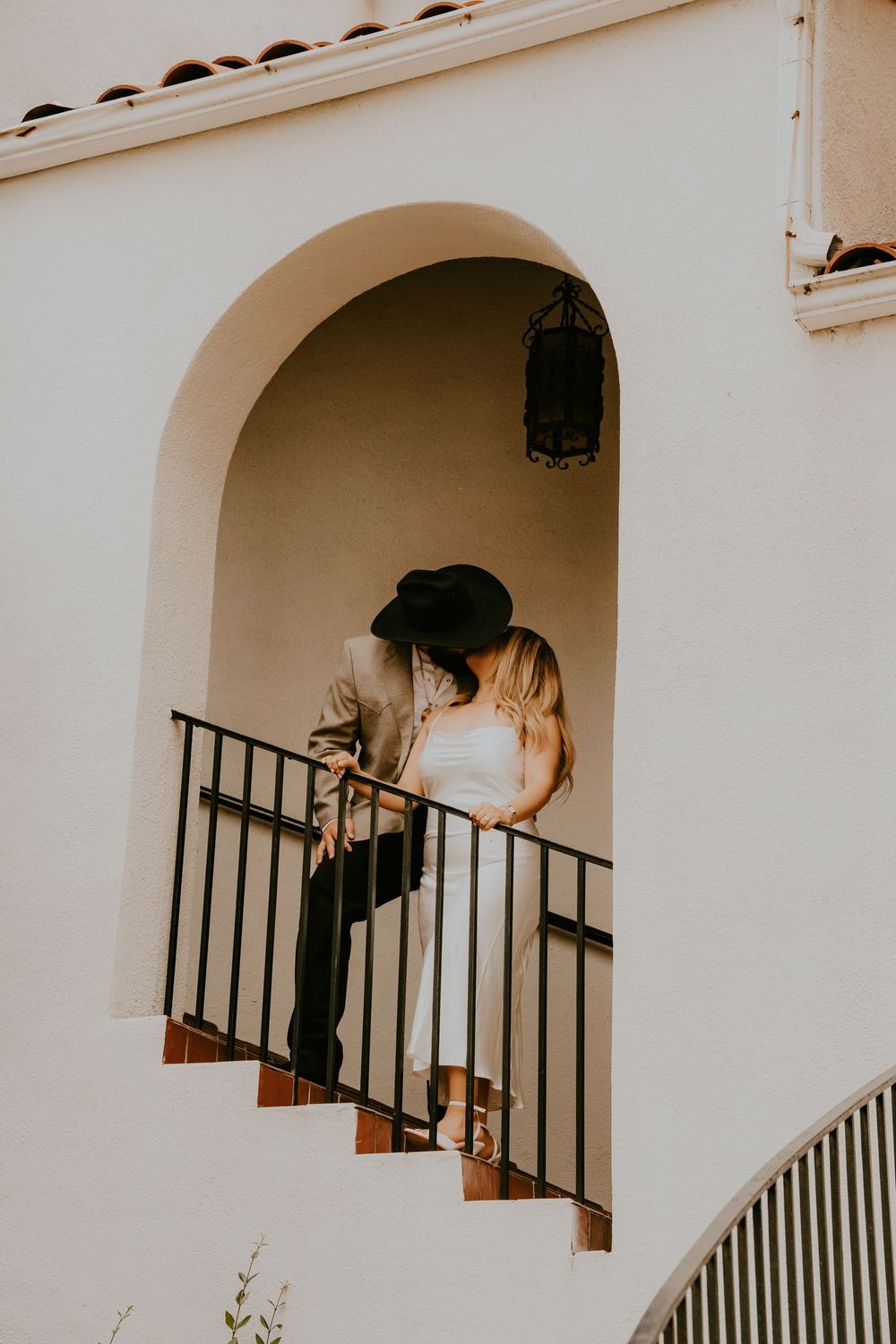 Romantic couple stands in white Mediterranean archway on exterior staircase during golden hour.