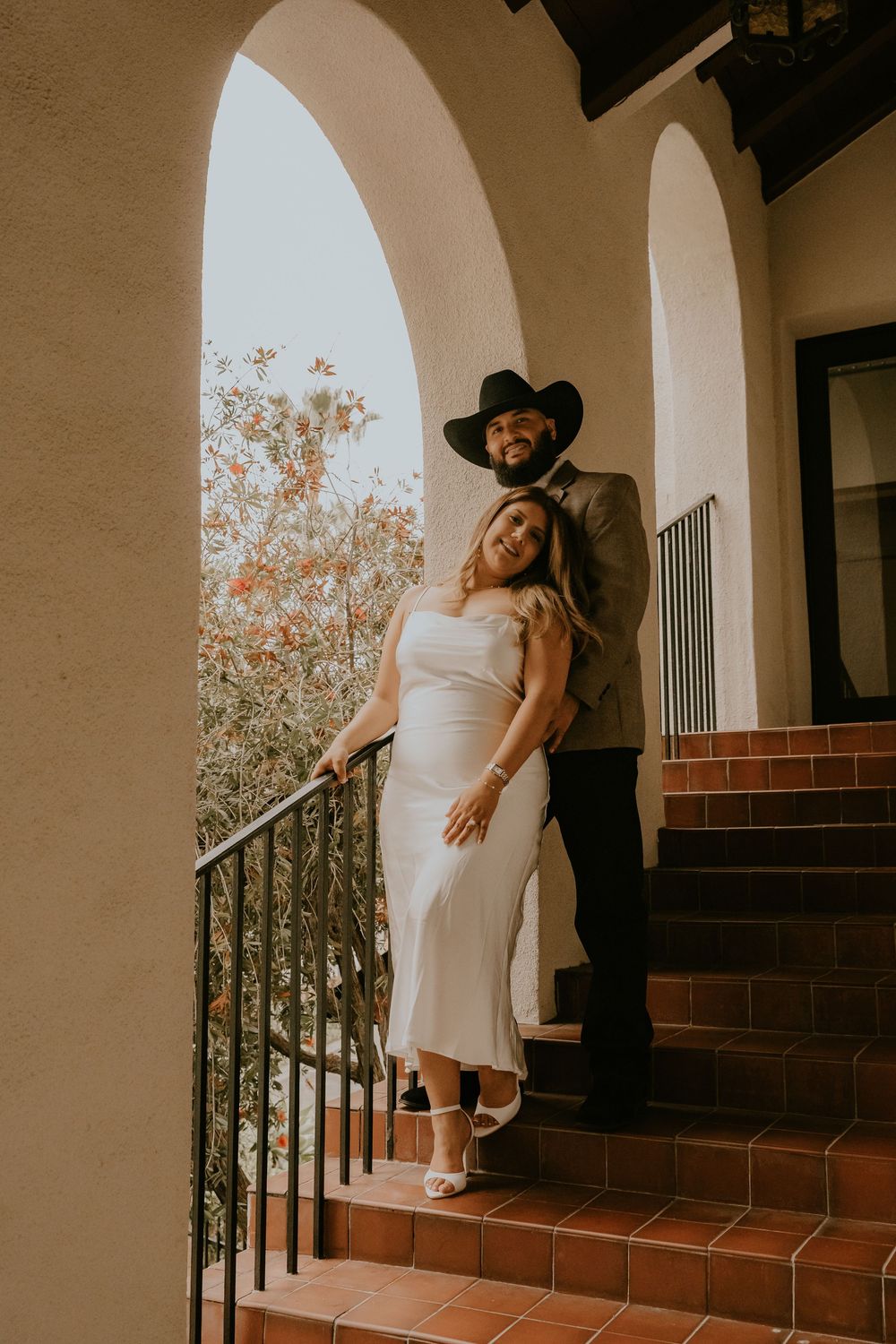 A romantic couple poses in an elegant white archway with brick stairs, creating a soft sepia-toned photograph.