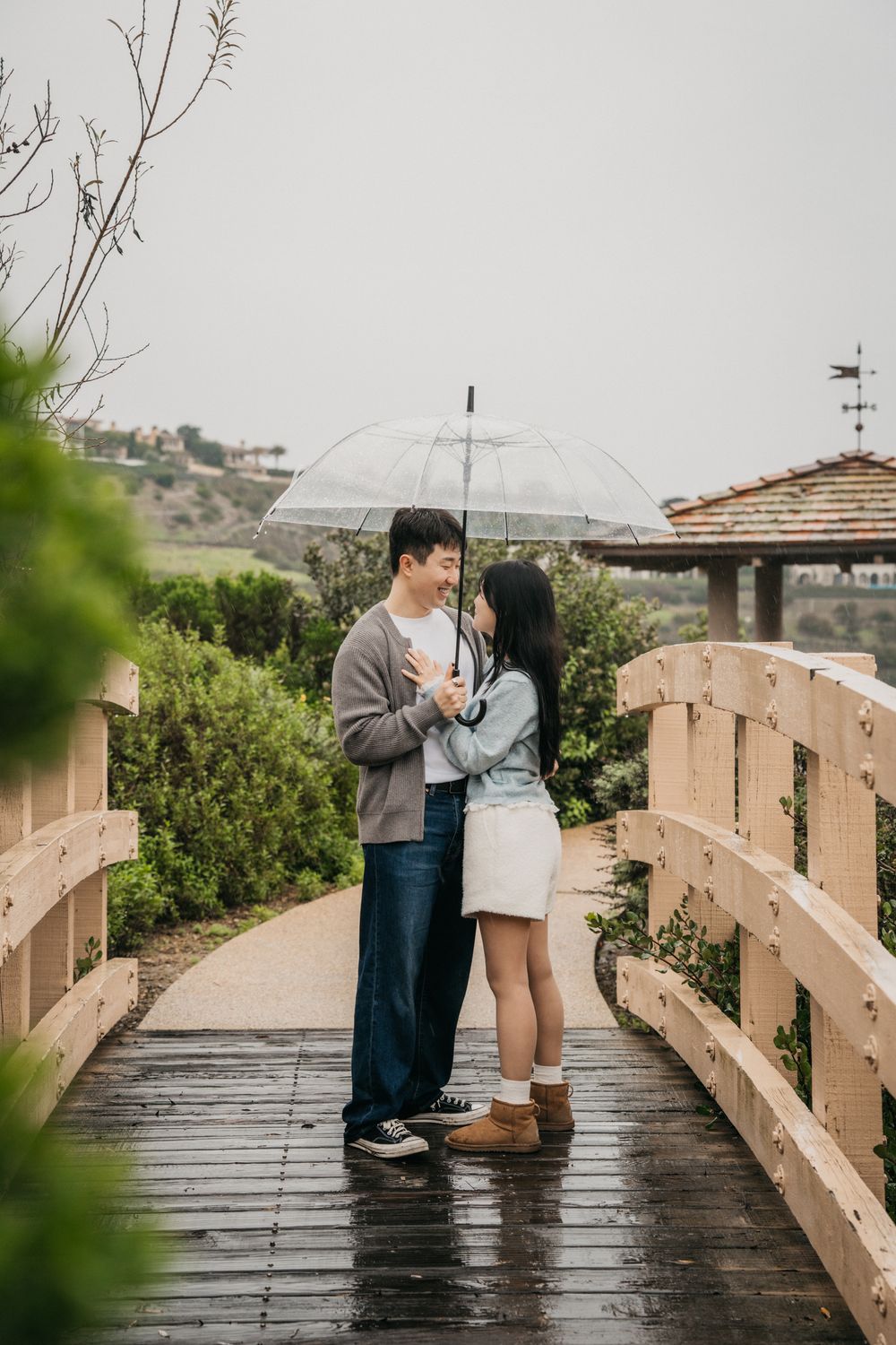 A couple shares a romantic moment under a clear umbrella on a wooden bridge during a rainy day.