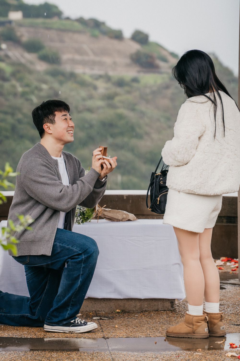 A romantic proposal moment captured outdoors on a hillside with rose petals scattered on the ground.