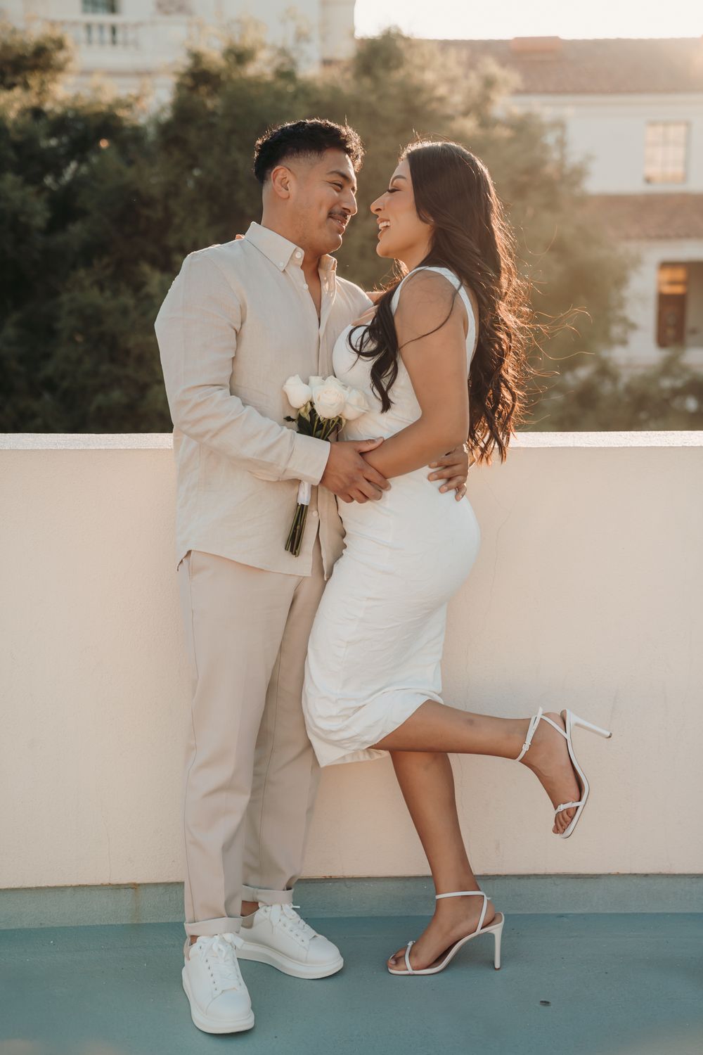 A romantic couple embraces on a sunny terrace, wearing matching white outfits with their bodies silhouetted against the sunset.