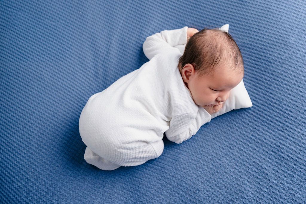 Newborn in white outfit lying on textured blue background during professional photo session.