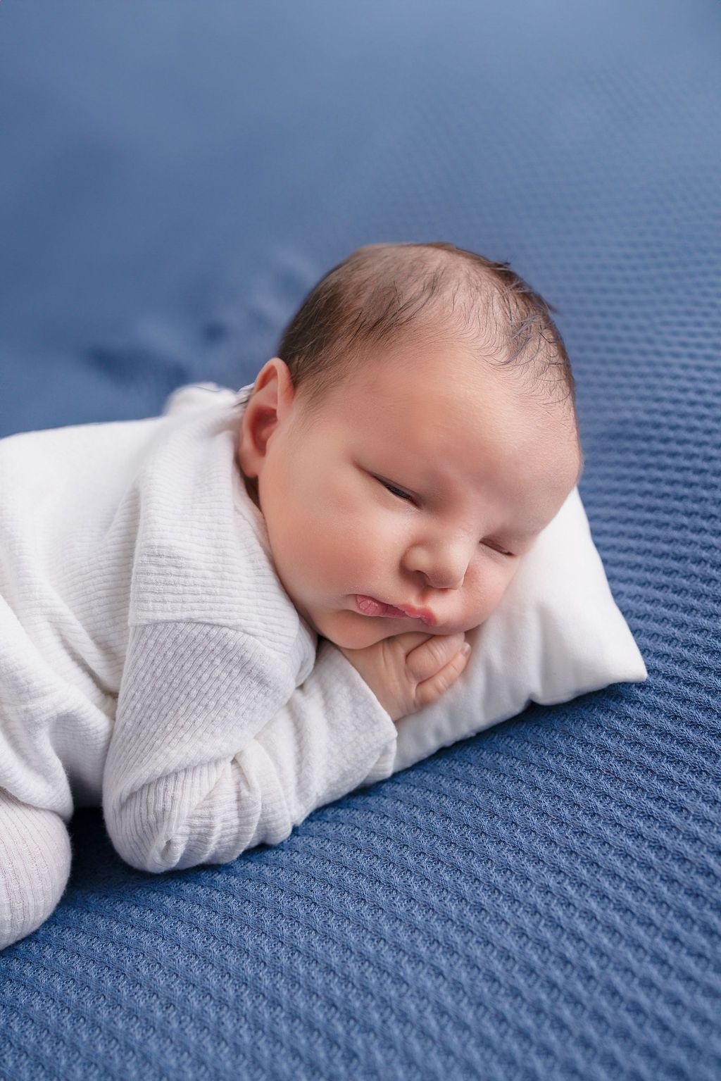 Sleeping newborn in white wrap positioned on blue textured fabric during portrait session.