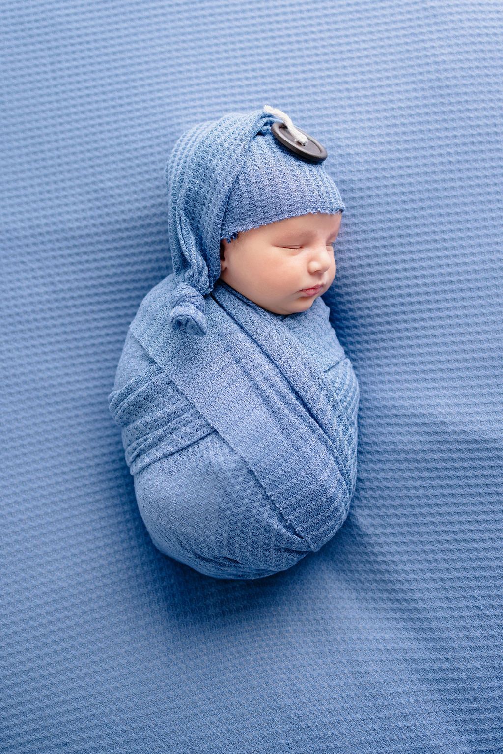 Overhead view of sleeping newborn swaddled in blue wrap on matching textured backdrop.
