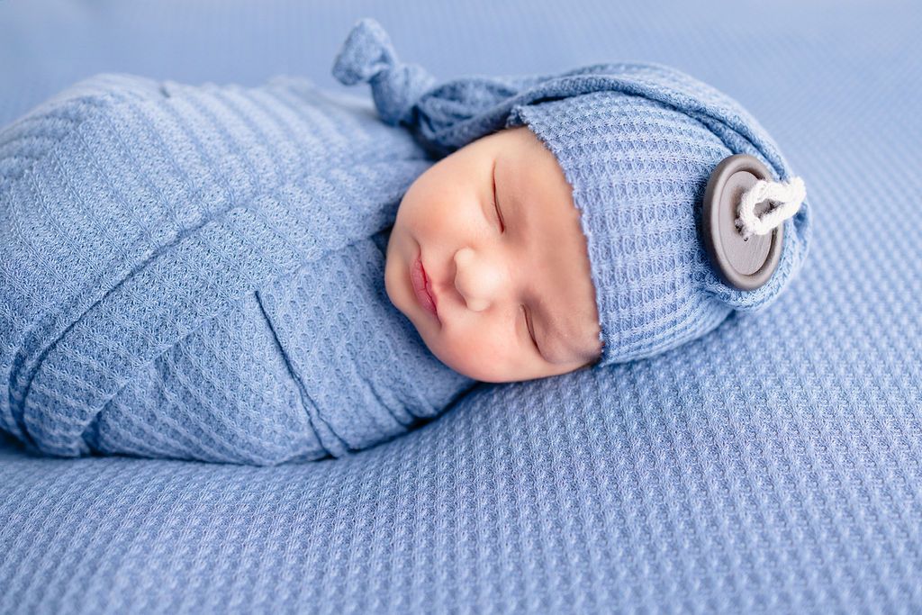 A sleeping newborn wrapped in soft blue fabric lies on a textured blue blanket in a gentle pose.